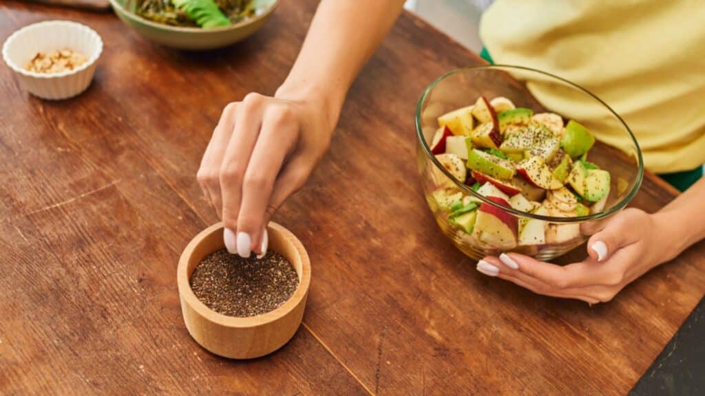 Cropped view of woman taking sesame seeds from wooden bowl near fresh fruit salad, vegetarian diet